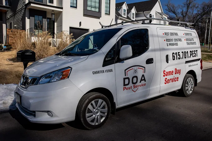 A white Nissan NV200 service van parked on a residential street in front of modern, two-story townhomes. The van is branded with the "DOA Pest Service" logo on the front door, "Same Day Service" in bold red text, and a phone number (615.701.PEST) on the side panel. Bullet points list their services, including pest control, termite treatment, and rodent control. A roof rack is mounted on top of the vehicle.