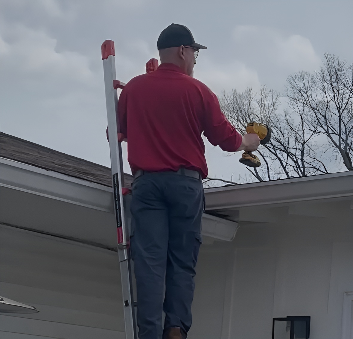 A technician viewed from behind, standing on a silver extension ladder to access the roofline of a white residential building. He is wearing a red long-sleeve shirt, grey cargo pants, and a dark baseball cap. He holds a yellow cordless power tool, likely a drill or a specialized dust applicator, while inspecting or treating the area near the gutters and roof edge under an overcast sky.